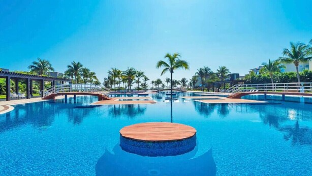 A large, clear swimming pool with a wooden deck in the center, surrounded by palm trees and a clear blue sky. - Casa Bonita, Oceanfront, Private pool condo, Mareazul (Playa del Carmen)