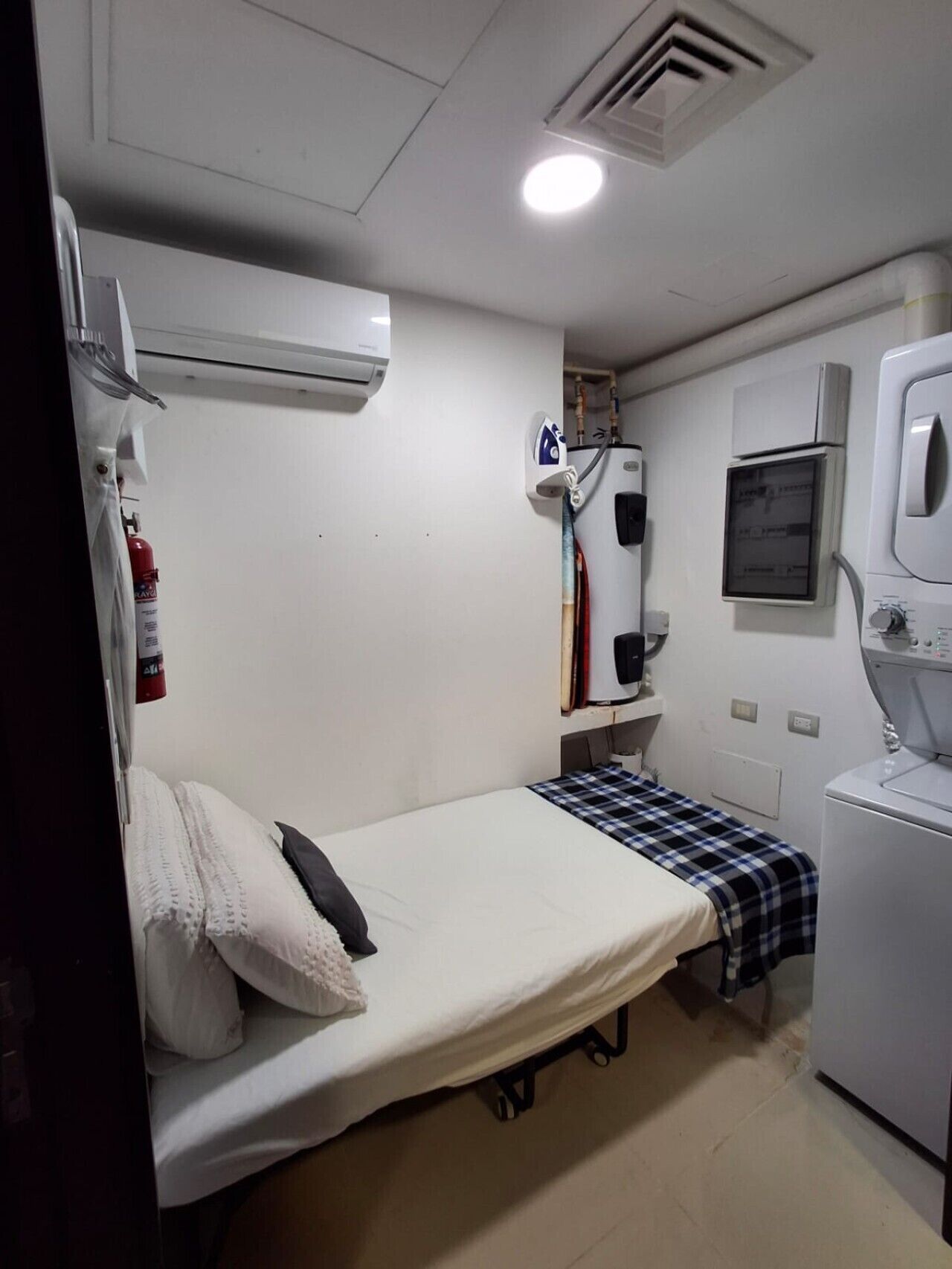 A small, compact room with a bed, a washing machine, a heater, and a small shelf with books.