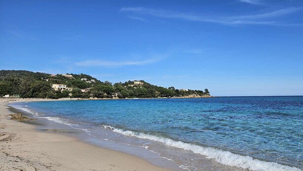 A beach with clear blue water, sandy shore, and buildings on a hillside in the background.
