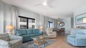 A living room with a blue sofa, a patterned armchair, a glass coffee table, and a dining area with a table and chairs. - The Palms 804 - RELAX AT THE  BEACH IN THIS GORGEOUS GULF FRONT CONDO! (Orange Beach)