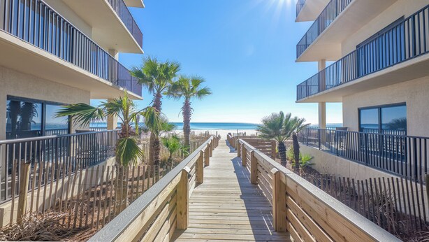 A wooden boardwalk leading to a beach, flanked by buildings with balconies and palm trees. - The Palms 804 - RELAX AT THE  BEACH IN THIS GORGEOUS GULF FRONT CONDO! (Orange Beach)