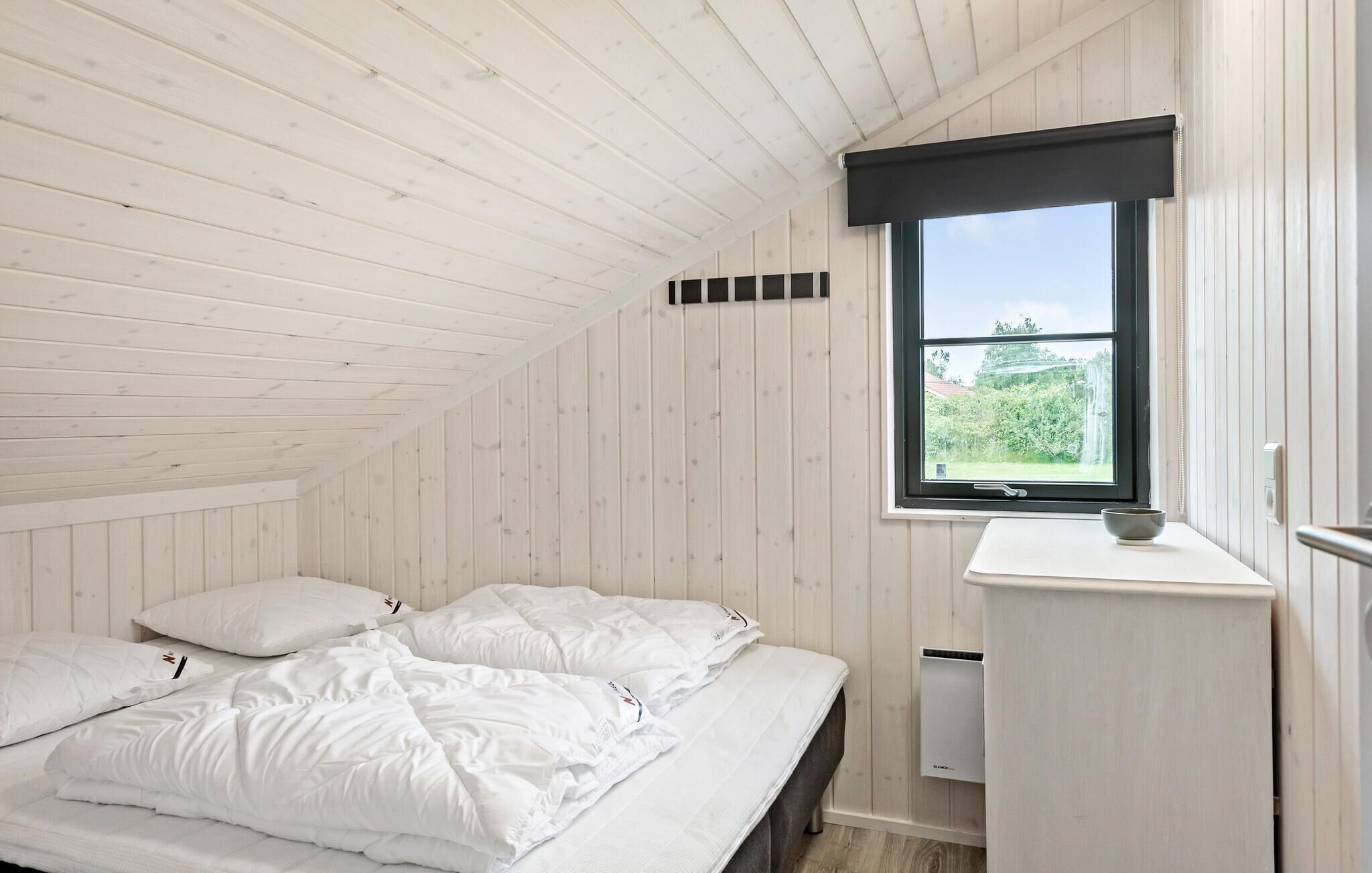A bedroom with a bed, white bedding, a window with a view of greenery, and a white wall with a black window frame.