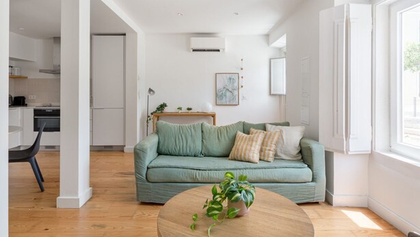 Ein modernes Wohnzimmer mit einem grünen Sofa, einem runden Holztisch mit einer Pflanze und einer Küche im Hintergrund.