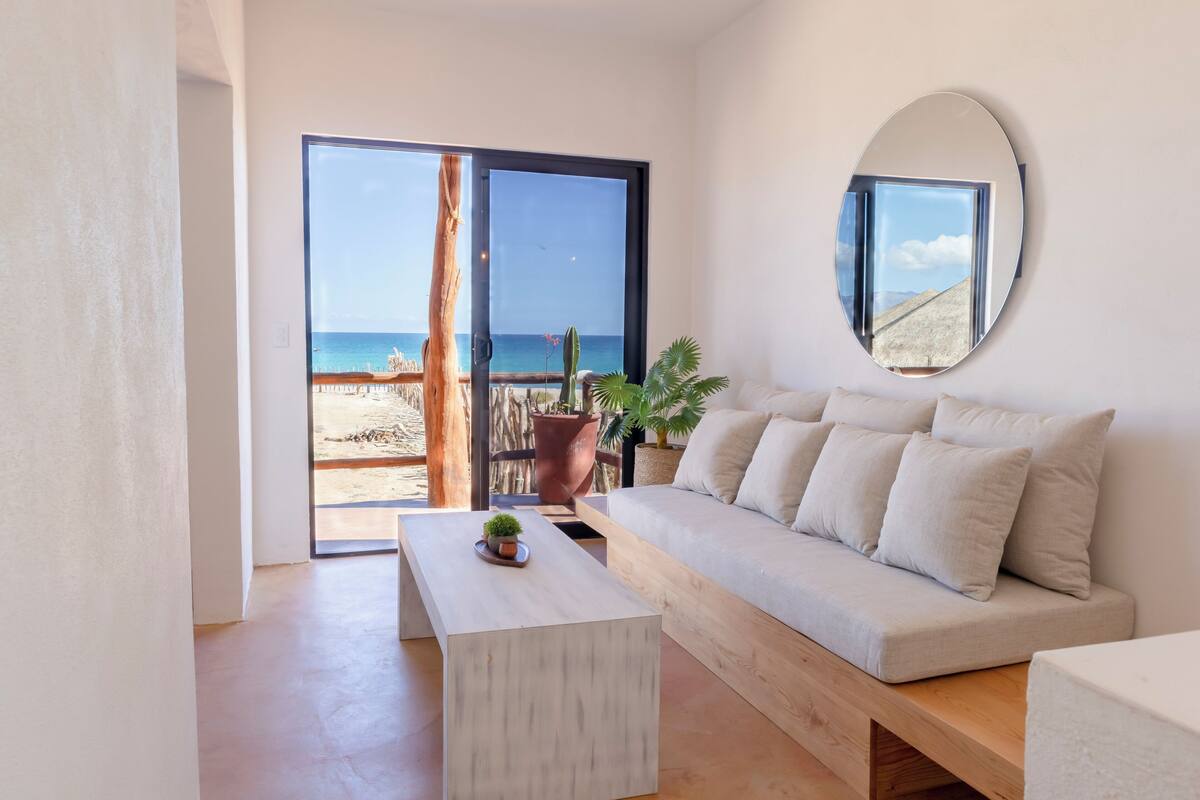 En moderne stue med sofa, sofabord og stort vindue med udsigt over en strand.