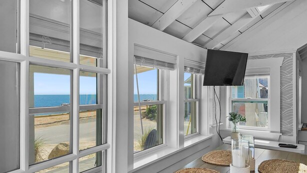 A spacious room with large windows offering ocean views, a flat-screen TV mounted on the wall, and woven mats on the floor. - Beachfront Escape in Dennis Port (Dennis Port)