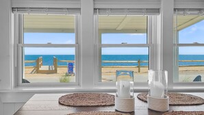 A room with three large windows overlooking a beach, featuring woven placemats and candle holders on a wooden table. - Beachfront Escape in Dennis Port (Dennis Port)