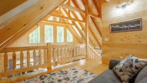 Um quarto de sótão acolhedor com teto de madeira, varanda com grades de madeira e uma janela grande que oferece vista para árvores.