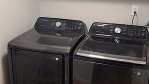 Une buanderie équipée de deux machines à laver et deux sèche-linge à chargement frontal, avec des placards blancs au-dessus et une prise de courant sur le mur.