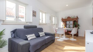 Un salon moderne avec un canapé gris, des chaises blanches, une table à manger en bois et un coin cuisine doté de meubles blancs et d’un meuble en bois orné de plantes.