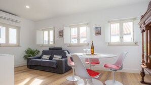 Un salon moderne avec un canapé gris, des chaises blanches et rouges, une petite table ronde sur laquelle se trouvent une bouteille et des verres, et de grandes fenêtres aux cadres blancs.