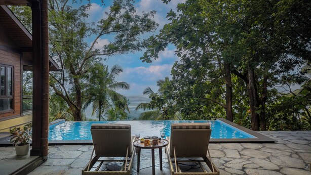 A poolside area with two wicker chairs and a small table, surrounded by lush greenery and a clear view of the ocean.