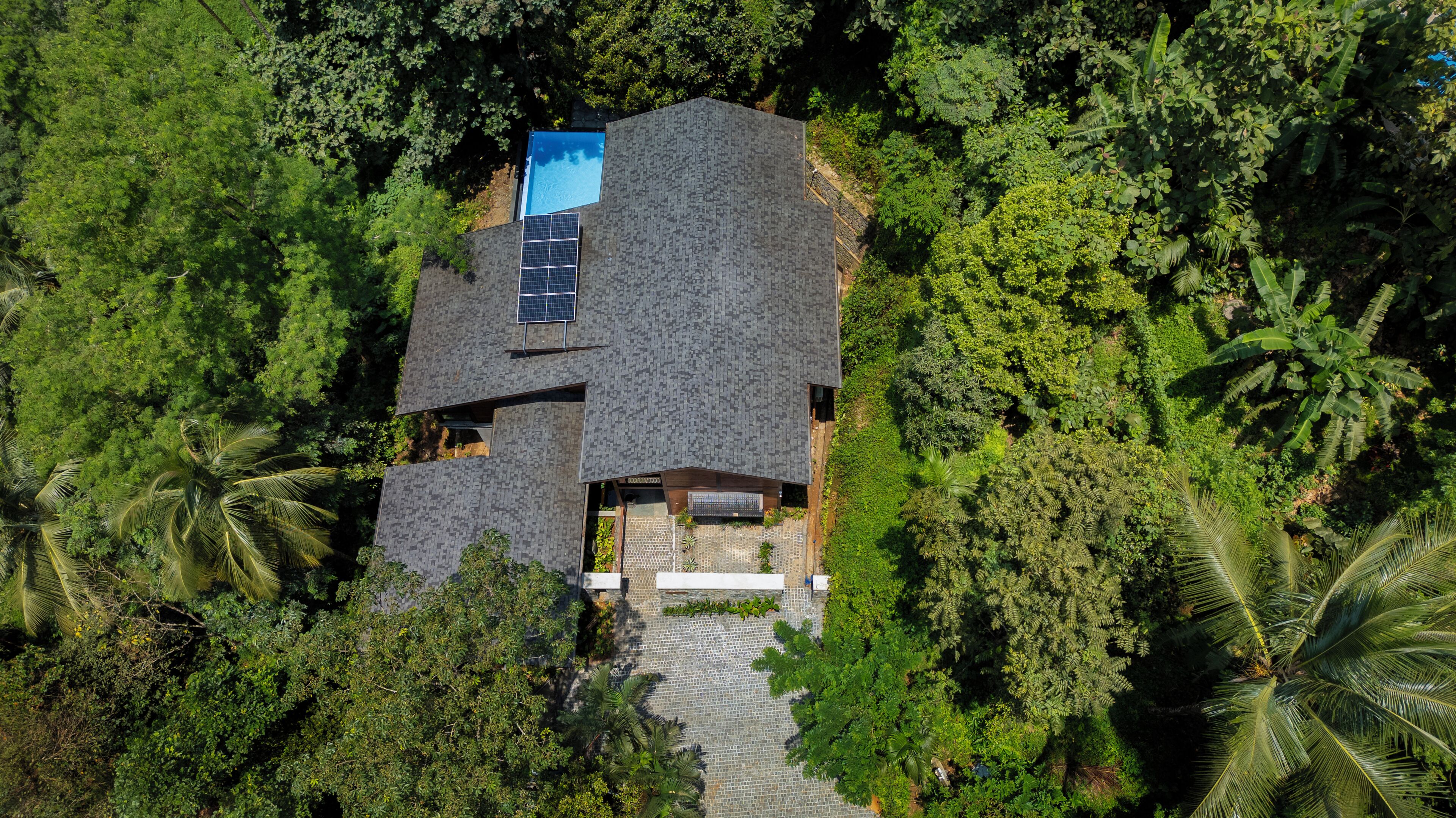 An aerial view of a house surrounded by dense tropical vegetation, featuring a solar panel roof and a visible swimming pool.