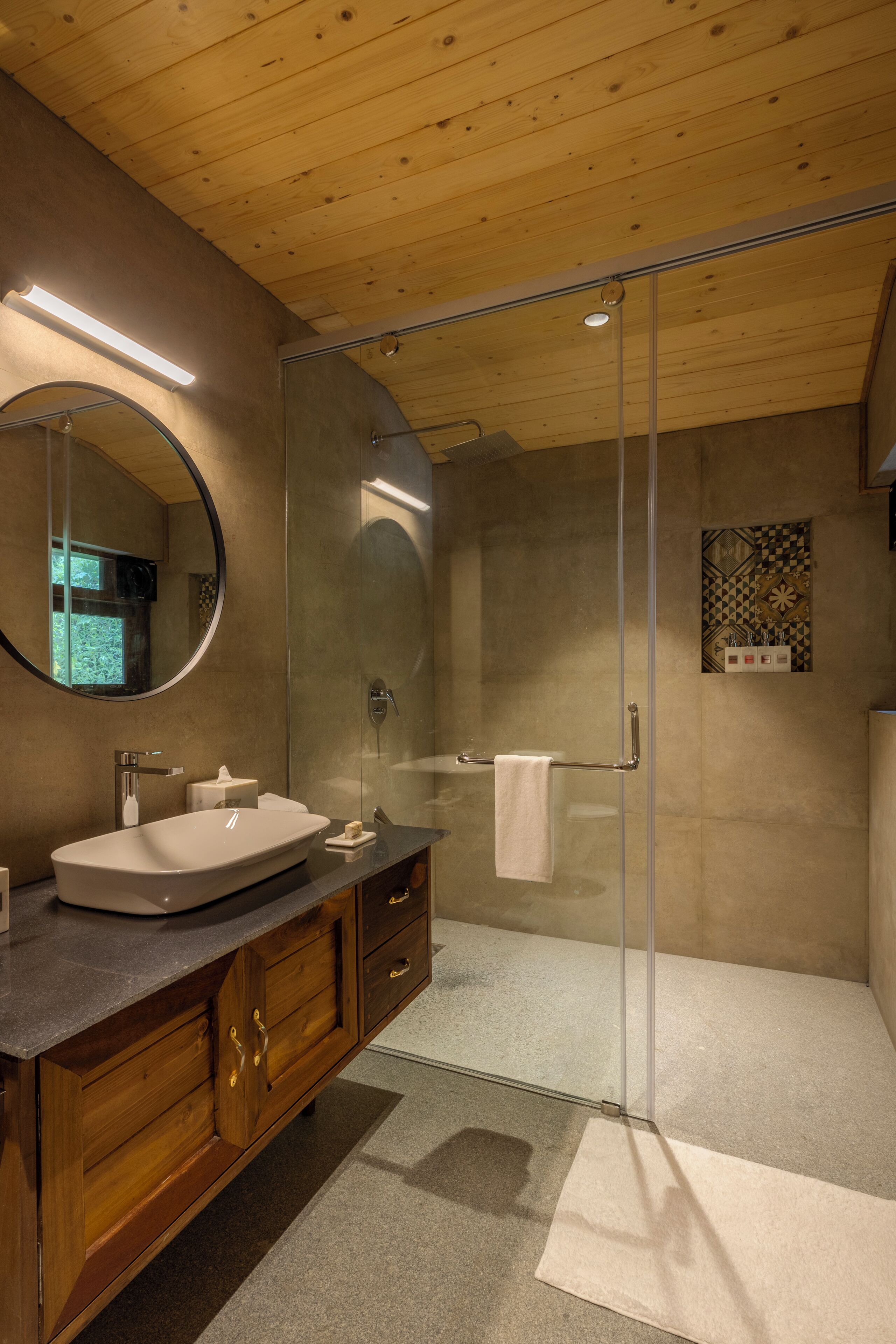 A modern bathroom with a glass-enclosed shower, a round mirror, and wooden cabinetry.