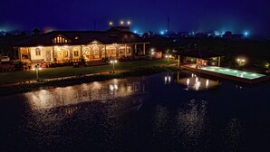 Hotel bem iluminado com piscina, área externa coberta e reflexo na água.