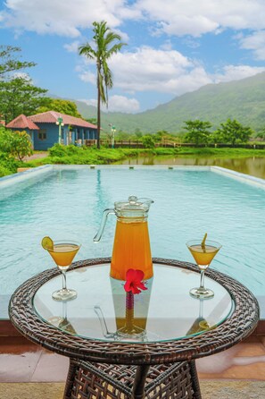 A glass table with a pitcher and two martini glasses, set by a pool with a mountain view.