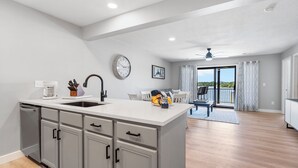 Cozinha moderna com armários cinzentos, bancada branca e lava-louças embutida. Há um espaço de jantar com mesa e cadeiras, e uma varanda com vista.