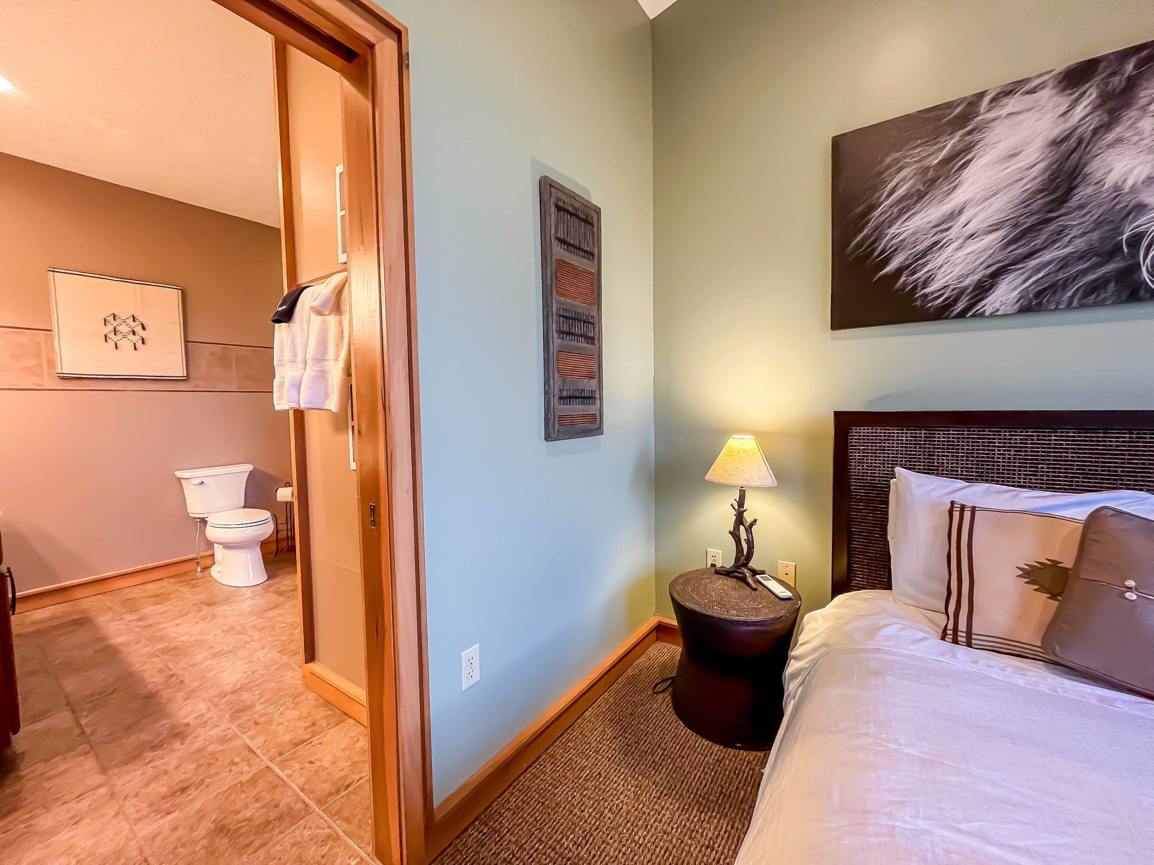 Una habitación de hotel con cama, una mesita de noche con lámpara, un cuadro en la pared y un baño visible a través de una puerta abierta.