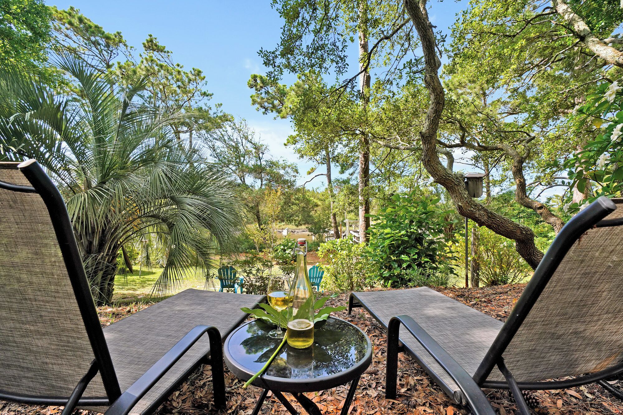 Deux fauteuils de jardin sont placés face à une table en verre sur laquelle se trouvent une bouteille et un verre. L’ensemble est entouré d’arbres et le ciel est dégagé.