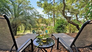 Twee terrasstoelen gericht op een glazen tafel met een fles en een glas, omgeven door bomen en een heldere lucht.