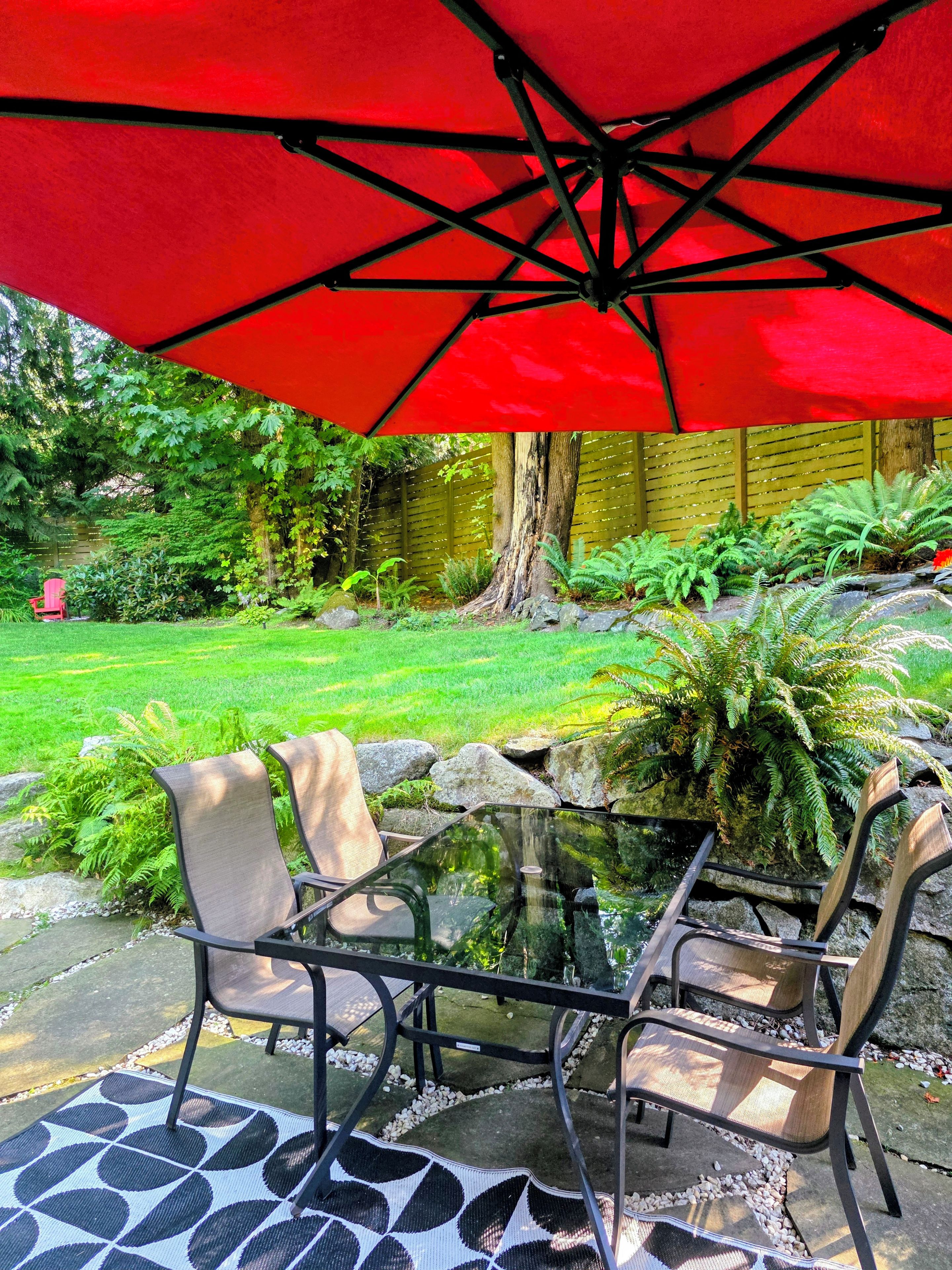 Un patio al aire libre con una mesa de vidrio, dos sillas y un paraguas rojo.