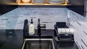Una cocina con encimera negra, un lavamanos, una cafetera, dispensador de jabón y escurreplatos. Hay estantes sobre la encimera con copas, tazas, cuencos y platos.