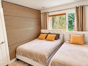 A bedroom with two beds, a wooden slat wall, a window with curtains, and a view of greenery outside.