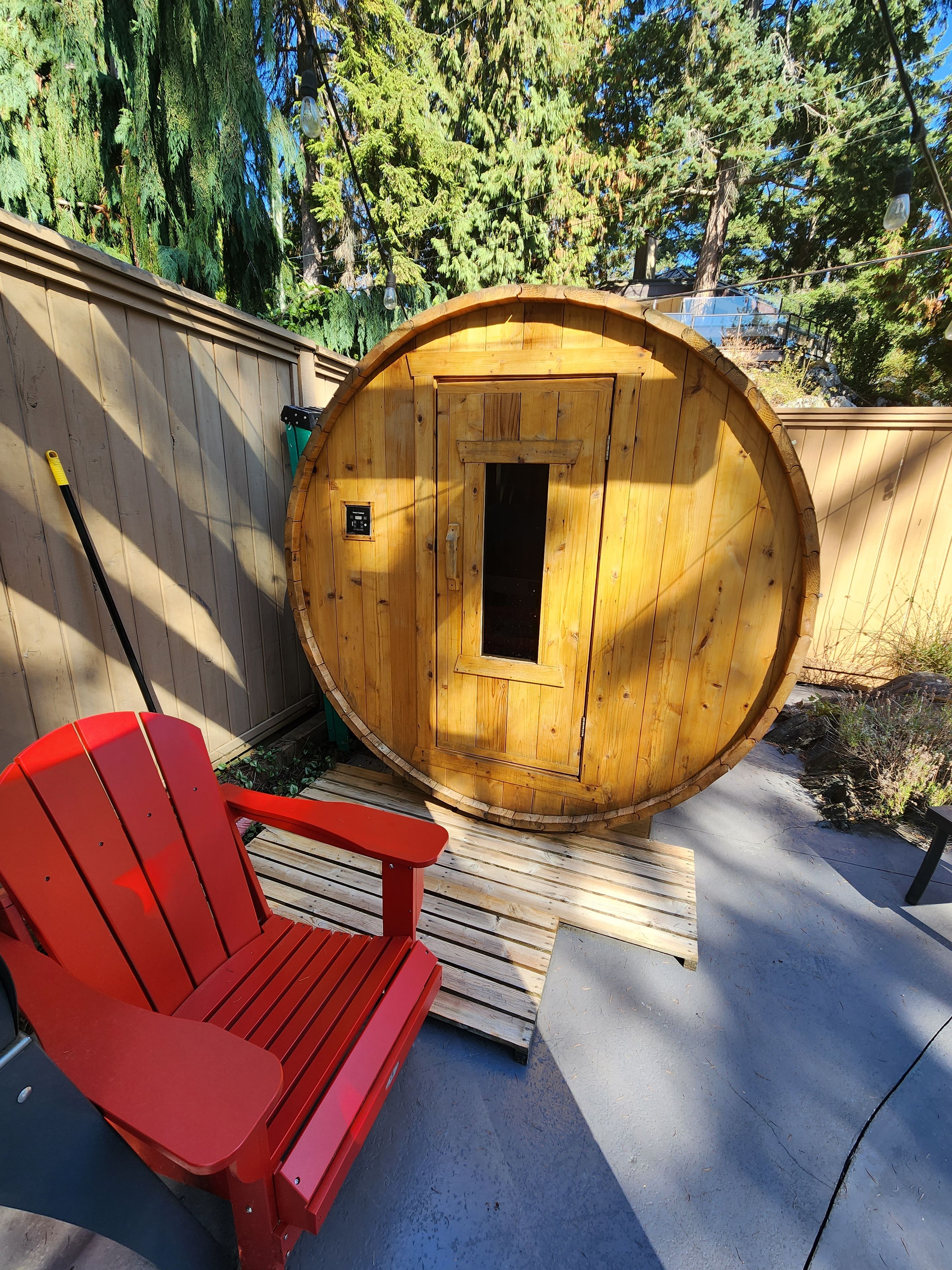 Un sauna en bois doté d’une porte circulaire, une chaise Adirondack rouge et une terrasse en bois.