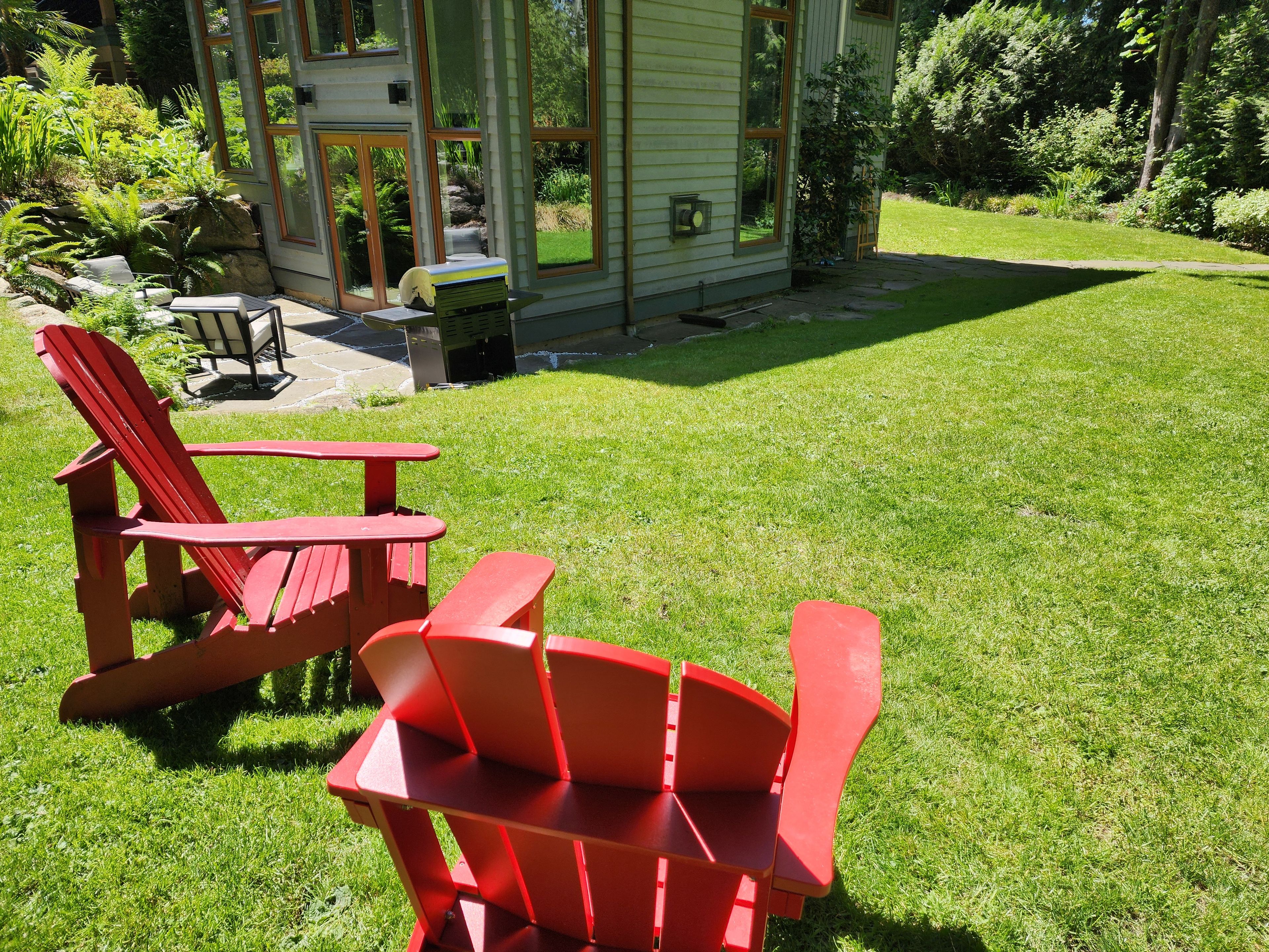 Un patio con sillas Adirondack rojas y una pequeña mesa, rodeado de un césped cuidado y una casa con ventanas grandes.