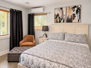 Quarto com cama, cadeira, mesa de cabeceira com abajur e duas janelas com cortinas.