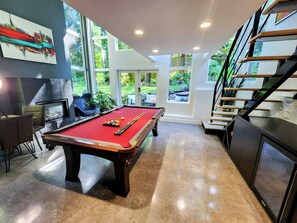 Een pooltafel met ballen, een bank en een stoel in een kamer met grote ramen en een trap.