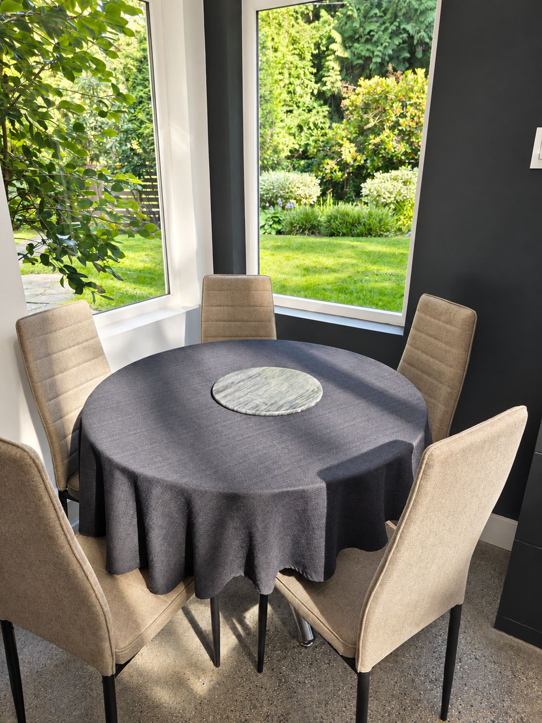 Une table ronde recouverte d’une nappe foncée, entourée de six chaises beiges, est placée devant de grandes fenêtres donnant sur un jardin.