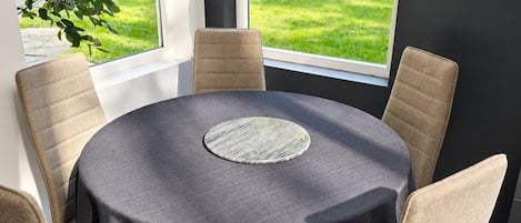 Une table ronde recouverte d’une nappe foncée, entourée de six chaises beiges, est placée devant de grandes fenêtres donnant sur un jardin.