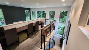 Una oficina en casa moderna con un escritorio, sillas y una estantería. Amplias ventanas ofrecen vistas al verdor exterior.