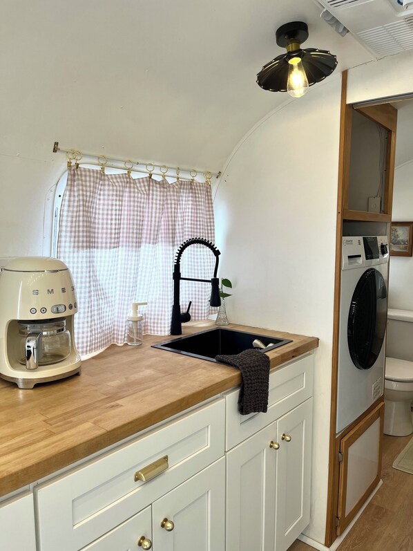 A compact kitchenette with a coffee maker, a sink, and a small window with a checkered curtain.