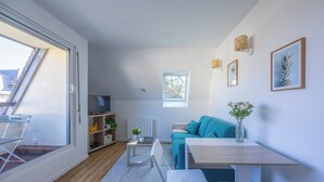 A modern living room with a sofa, a coffee table, a TV, and a dining table. There is a window with a view of the outside, a radiator, and a shelf with decorative items. - Duplex Les Mouettes - Welkeys (Cabourg)