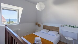 A bedroom with a bed, a window, a white radiator, and a view of the sea. - Duplex Les Mouettes - Welkeys (Cabourg)