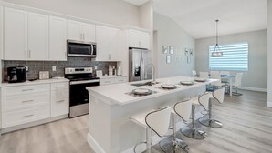 Una cocina moderna con armarios blancos, una isla central con taburetes altos, electrodomésticos de acero inoxidable y un amplio ventanal con persianas.