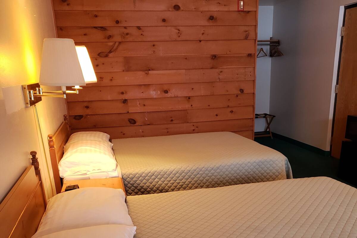 A room with two beds, wooden panel walls, a wall-mounted lamp, and a coat rack.