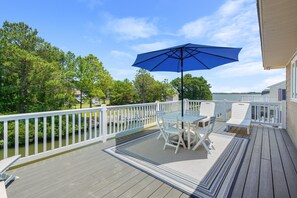 A wooden deck with a table and chairs, a blue umbrella, and a white railing overlooking a body of water. - Bayside Bliss | Chincoteague Waterfront w/ Dock, Enclosed Porch Dining, Sunset Views & 4BR Comfort (Chincoteague Island)