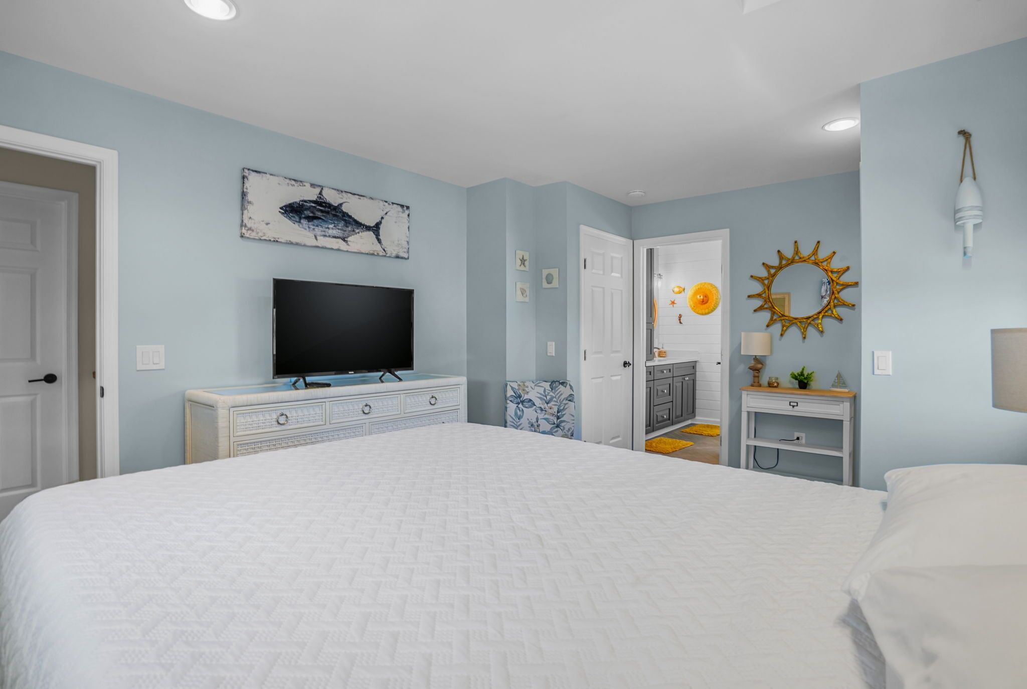 Une chambre avec un grand lit, un téléviseur posé sur une commode, une porte donnant sur une autre pièce et un miroir en forme de soleil accroché au mur.