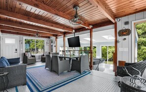 A covered outdoor area with a dining table and chairs, a TV mounted on the ceiling, and a view of the outdoors. - Bayside Bliss | Chincoteague Waterfront w/ Dock, Enclosed Porch Dining, Sunset Views & 4BR Comfort (Chincoteague Island)