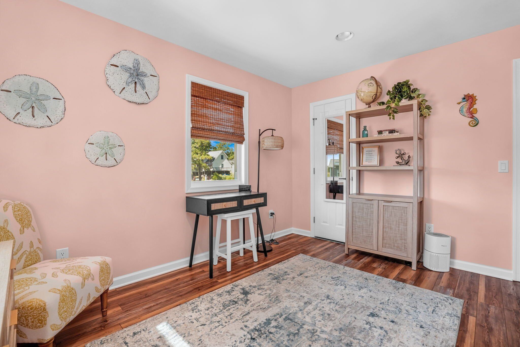 Una habitación con paredes rosas, suelo de madera, un escritorio con computadora, una silla, una estantería y una ventana con persianas.