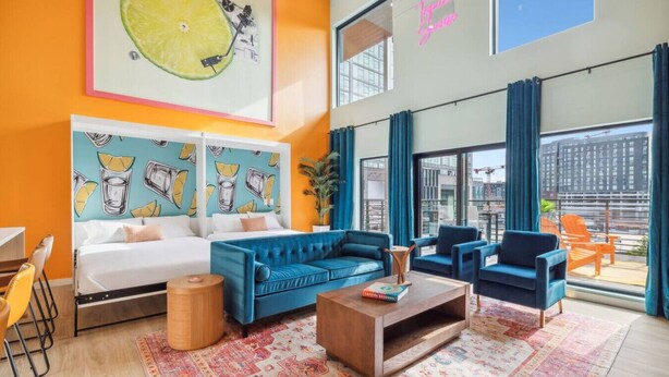 A modern hotel room with a large bed, a blue sofa, a coffee table, and a city view through large windows. - Tequila Sunrise Downtown Condo, Walk to Broadway! (Nashville)