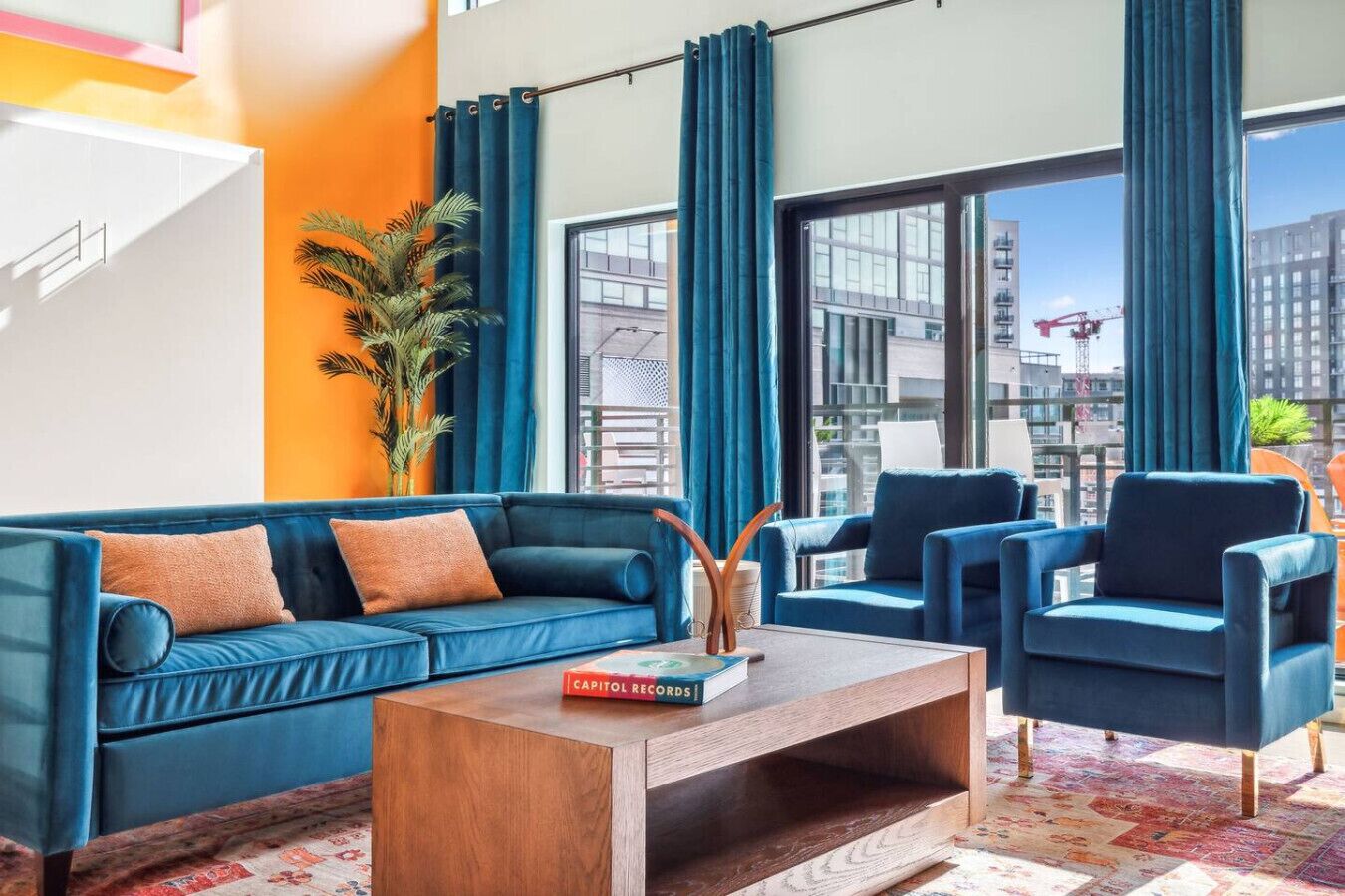 A modern living room with a blue sofa, matching armchairs, and a wooden coffee table. A large window offers a view of the cityscape.