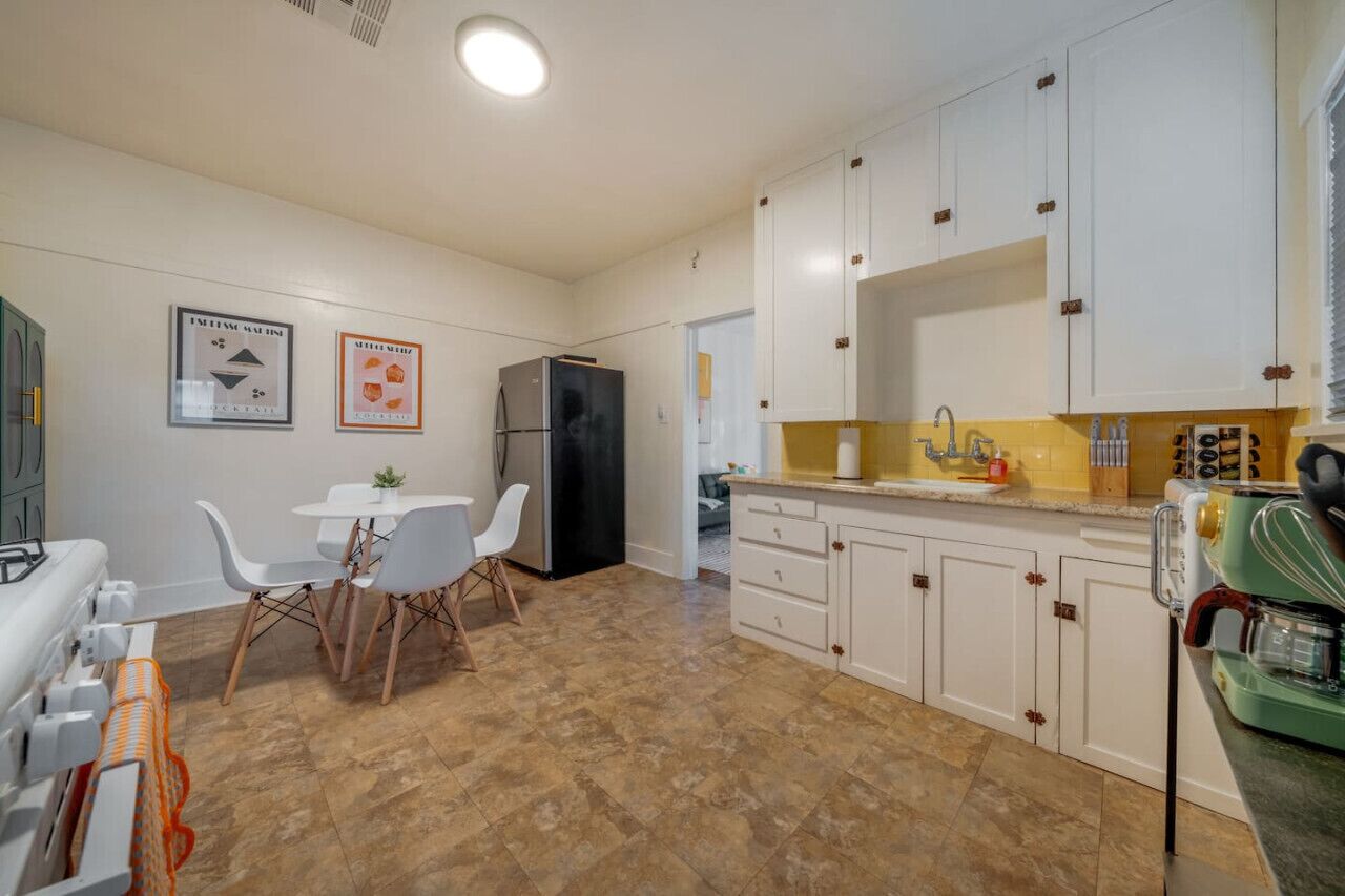 A compact kitchen with white cabinets, a black refrigerator, a dining table with chairs, and a coffee machine.