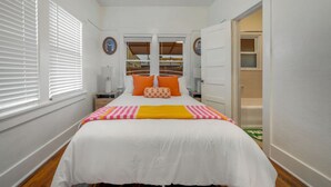 A bedroom with a bed, white walls, a window with blinds, a door leading to a bathroom, and a small table with a lamp. - Close to UofA / 1 BR 1 BA / Unique / Patio & Porch (Tucson)
