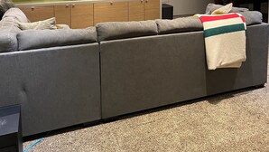 Un salon moderne avec un canapé d’angle gris, un téléviseur à écran plat et un coin cuisine doté de placards et d’une fenêtre.