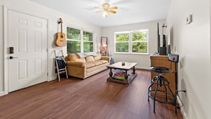 A living room with a wooden floor, a beige sofa, a coffee table, a guitar, and a ceiling fan. - Lake Cabin, 10 minutes from Callaway Gardens  (West Point)