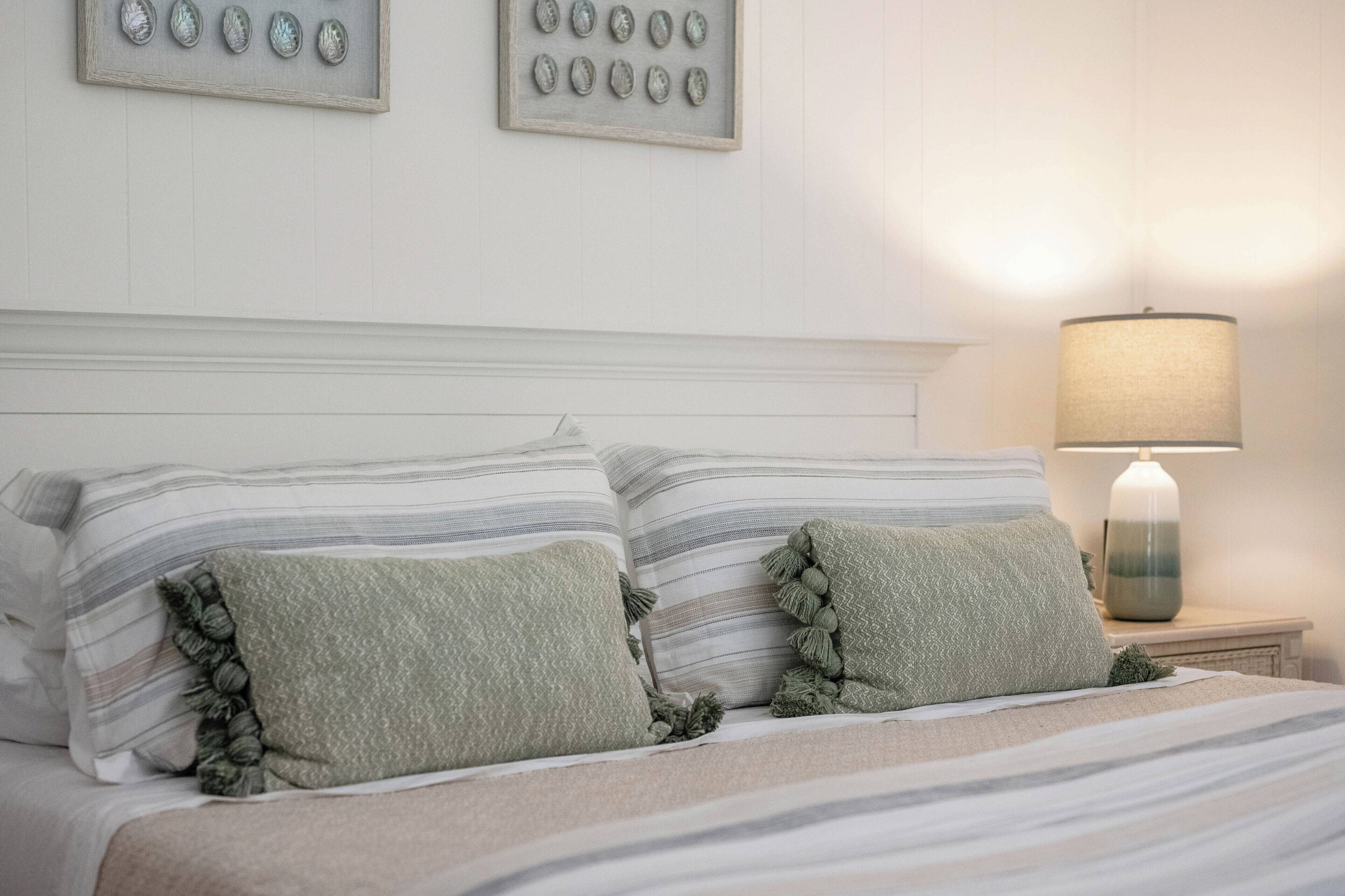 Una cama bien tendida con ropa de cama a rayas y dos almohadas, una lámpara de noche y dos elementos decorativos enmarcados en la pared.