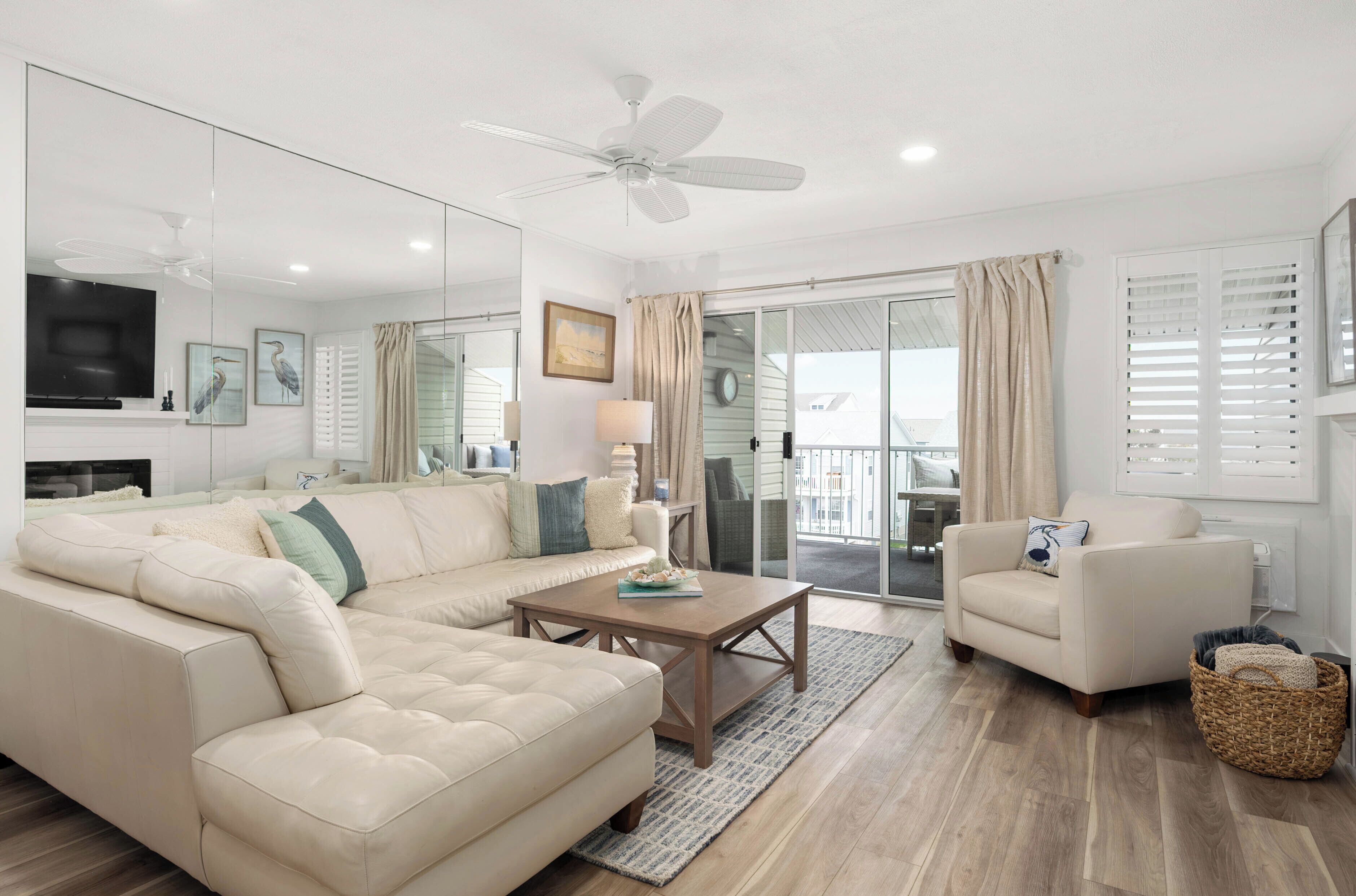 A modern living room with a beige sofa, a wooden coffee table, and a white armchair. There is a ceiling fan, a TV mounted on the wall, and a large window with curtains.